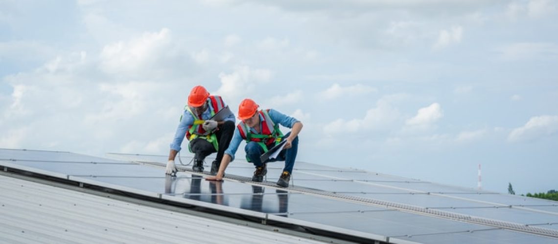 Technician wearing safety harness belt during installing the solar panels on roof structure of building factory,Economic energy and cost saving concept.